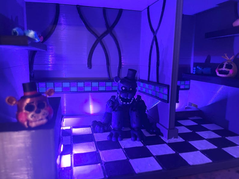 Op de afbeelding: Een diorama met een donker, verweerd berenfiguur met een hoge hoed op een zwart-wit geblokte vloer. Een kleinere berenkop met een hoge hoed staat op een richel aan de linkerkant en een gele vogelachtige kop staat op een plank aan de rechterkant.