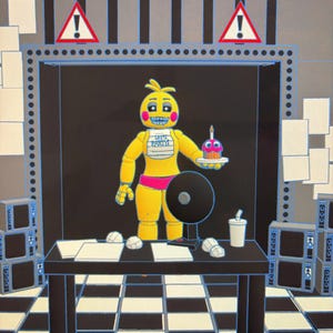May include: A yellow animatronic chicken figure with pink cheeks and a white bib reading LET'S PARTY! holds a cupcake with a candle. The figure stands behind a black desk with papers, a fan, and a cup, set against a black and white checkered floor.