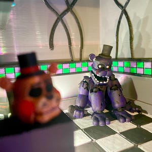 Op de afbeelding: Een paars berenfiguurtje met een hoge hoed en een microfoon zittend op een zwart-wit geblokte vloer. Op de voorgrond is een wazig oranje berenfiguurtje met een zwarte hoge hoed en rode accenten gedeeltelijk zichtbaar in het frame.
