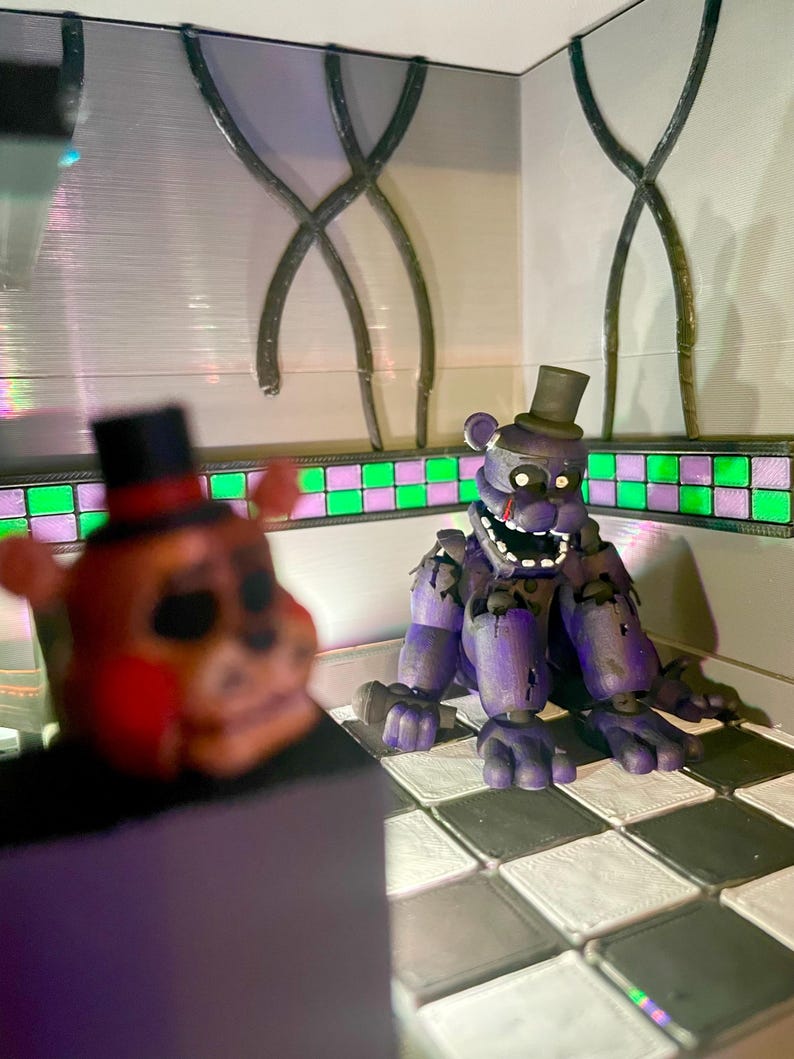 Op de afbeelding: Een paars berenfiguurtje met een hoge hoed en een microfoon zittend op een zwart-wit geblokte vloer. Op de voorgrond is een wazig oranje berenfiguurtje met een zwarte hoge hoed en rode accenten gedeeltelijk zichtbaar in het frame.