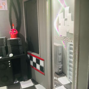 K&ouml;nnte beinhalten: Ein Miniatur-Diorama-Raum mit schwarz-wei&szlig; kariertem Boden, einer schwarzen Konsole mit einer rosa Cupcake-Figur und einem Durchgang zu einem Nebenraum mit wei&szlig;en Wandpaneelen und einem hohen grauen Schrank, alles in einer strukturierten grauen Optik.