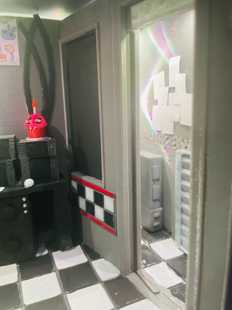 K&ouml;nnte beinhalten: Ein Miniatur-Diorama-Raum mit schwarz-wei&szlig; kariertem Boden, einer schwarzen Konsole mit einer rosa Cupcake-Figur und einem Durchgang zu einem Nebenraum mit wei&szlig;en Wandpaneelen und einem hohen grauen Schrank, alles in einer strukturierten grauen Optik.