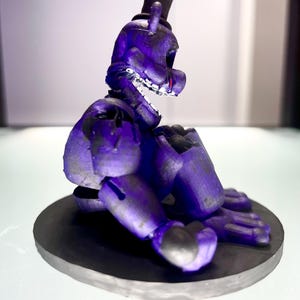Puede incluir: Figura morada de un oso con sombrero de copa negro, sentado sobre una base circular negra. La figura tiene dientes visibles y detalles rojos. El dise&ntilde;o sugiere un personaje de videojuego o serie animada.