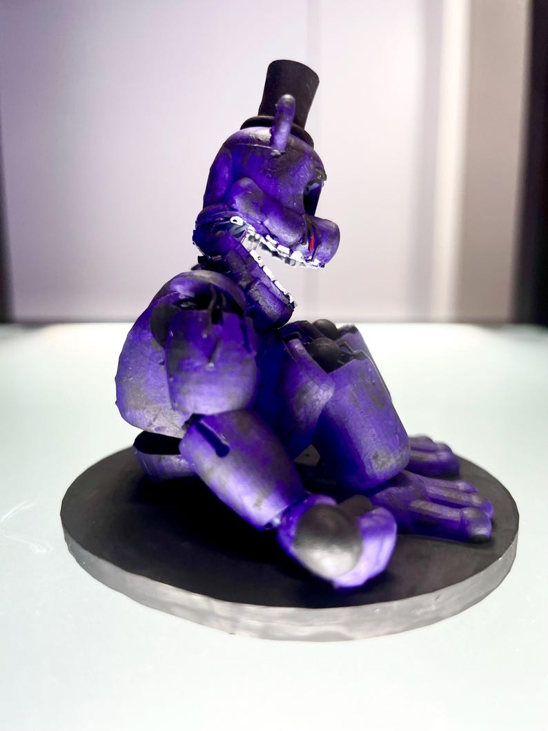 Puede incluir: Figura morada de un oso con sombrero de copa negro, sentado sobre una base circular negra. La figura tiene dientes visibles y detalles rojos. El dise&ntilde;o sugiere un personaje de videojuego o serie animada.