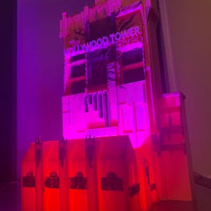 Op de afbeelding: Een model van het Hollywood Tower Hotel, verlicht met rode en paarse lichten. De gevel van het gebouw is gedetailleerd, met de woorden "Hollywood Tower Hotel" zichtbaar. Het model is een miniatuurreplica van de iconische structuur.
