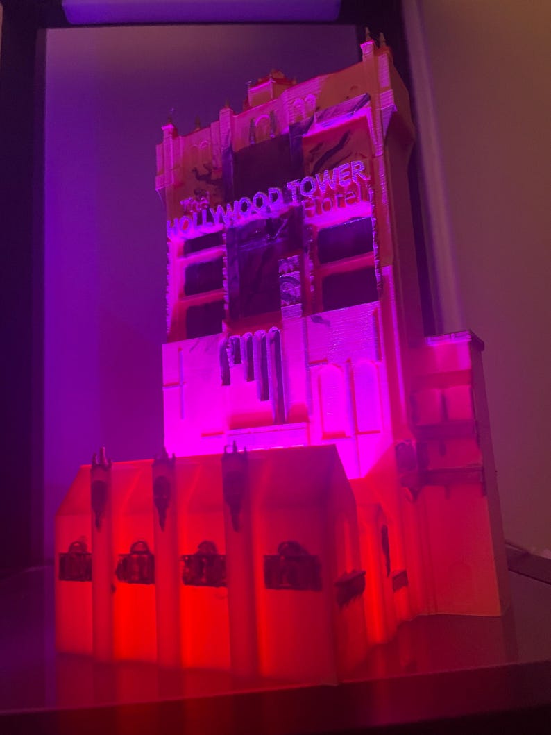 Op de afbeelding: Een model van het Hollywood Tower Hotel, verlicht met rode en paarse lichten. De gevel van het gebouw is gedetailleerd, met de woorden "Hollywood Tower Hotel" zichtbaar. Het model is een miniatuurreplica van de iconische structuur.