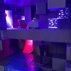 Puede incluir: Un diorama impreso en 3D con tem&aacute;tica de Minecraft con un esquema de iluminaci&oacute;n morado y azul. Una peque&ntilde;a figura de un pollo montando un carro de minas est&aacute; en el nivel superior. Una figura de un creeper negro est&aacute; en el nivel inferior. El diorama est&aacute; iluminado desde el interior con luces rojas y azules.