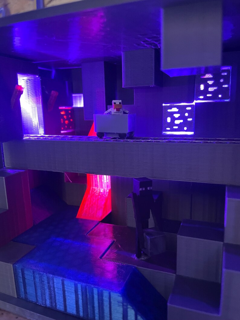 Puede incluir: Un diorama impreso en 3D con tem&aacute;tica de Minecraft con un esquema de iluminaci&oacute;n morado y azul. Una peque&ntilde;a figura de un pollo montando un carro de minas est&aacute; en el nivel superior. Una figura de un creeper negro est&aacute; en el nivel inferior. El diorama est&aacute; iluminado desde el interior con luces rojas y azules.