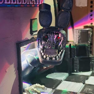 May include: A 3D printed figurine of a damaged blue rabbit character with long ears, exposed mechanical inner workings, and glowing red eyes. The figure is emerging from a doorway next to a sign that reads CELEBRATE, set on a chequered floor.