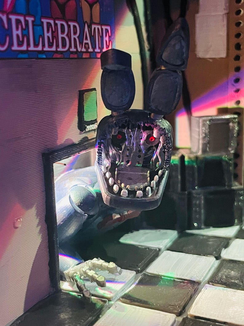May include: A 3D printed figurine of a damaged blue rabbit character with long ears, exposed mechanical inner workings, and glowing red eyes. The figure is emerging from a doorway next to a sign that reads CELEBRATE, set on a chequered floor.
