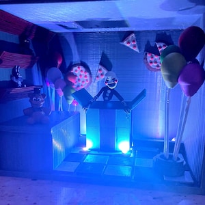 Puede incluir: Un diorama con personajes y elementos de la serie de videojuegos Five Nights at Freddy's. La escena incluye una marioneta que emerge de una caja, globos, porciones de pizza y otras figuras, todo bajo iluminaci&oacute;n azul.