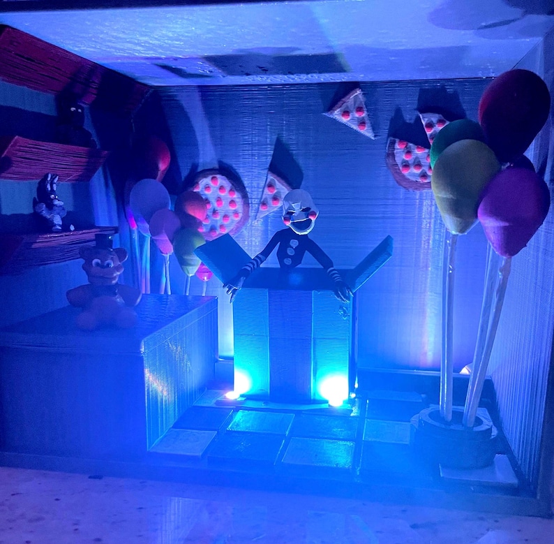 Puede incluir: Un diorama con personajes y elementos de la serie de videojuegos Five Nights at Freddy's. La escena incluye una marioneta que emerge de una caja, globos, porciones de pizza y otras figuras, todo bajo iluminaci&oacute;n azul.
