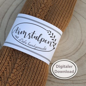 May include: A rolled-up, knitted brown arm warmer with a white label wrapped around it. The label reads "Armstulpen mit Liebe handgemacht" in script. A small, oval sticker with "Digitaler Download" is also visible.