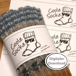 Op de afbeelding: Handgebreide sokken met een label met de tekst "Coole Socke" en een cartoon sok met een zonnebril. De sokken zijn gemaakt van blauw, grijs en bruin garen. Een sticker zegt "Digitaler Download".