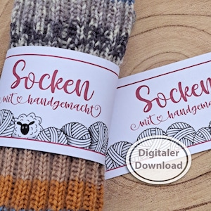 May include: A pair of colorful socks with a label that reads "Socken mit handgemacht" in burgundy script. The label features illustrations of yarn balls and a sheep. A digital download badge is also visible.