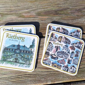 Könnte beinhalten: Quadratische Untersetzer mit dem Text "Rietberg an der Ems" und Abbildungen von Gebäuden. Ein Untersetzer zeigt eine detaillierte Illustration eines Gebäudes mit Turm. Der andere hat eine Collage verschiedener Gebäude.