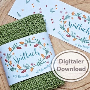 May include: A green knitted washcloth with a decorative label wrapped around it. The label features the word "Spültuch" in a decorative oval with floral and star accents. The label also includes the text "100% Baumwolle" and "mit liebe handgemacht".