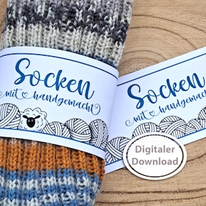 Op de afbeelding: Close-up van een paar handgemaakte sokken met een label met de tekst "Socken mit handgemacht" in blauw schrift. De sokken zijn een mix van grijs, blauw en oranje garen. Het label bevat illustraties van schapen en bollen wol.