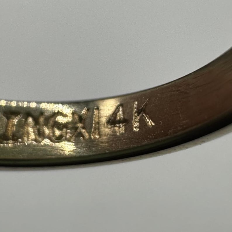 May include: Close-up of a gold ring with the inscription "INGAI 14K" engraved on the inside. The ring is a warm gold color and appears to be well-worn.
