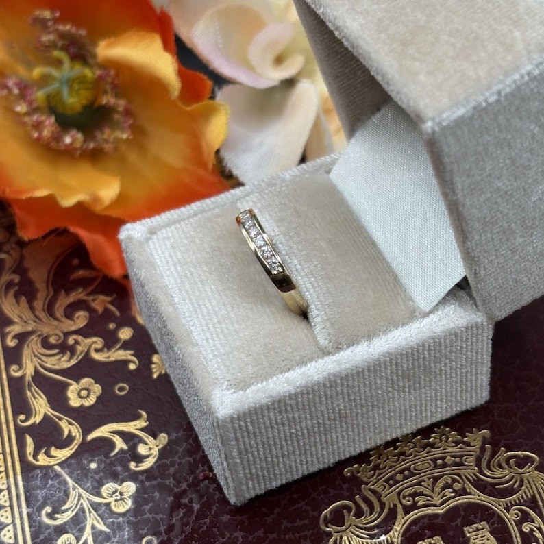 May include: A gold band ring with small, clear stones set across the top, displayed in a gray velvet ring box. The ring is set against a backdrop of flowers and a decorative book.