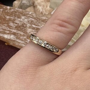 May include: A gold band ring with a row of small, clear gemstones set across the top. The ring is worn on a finger, showcasing its polished, reflective surface. The ring's design is simple and elegant.
