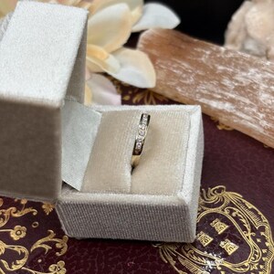 May include: A gold band ring with small, clear gemstones set in a row. The ring is displayed inside a light gray velvet ring box. The box is open, revealing the ring against a soft, neutral background.