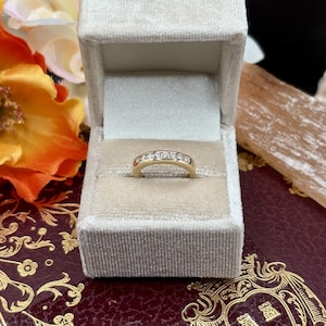 May include: A gold band ring with a row of small, clear gemstones set across the top. The ring is displayed inside a light beige velvet ring box with a hinged lid. The background includes floral elements and a patterned surface.