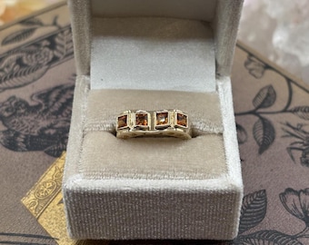 May include: A gold ring with four square, amber-colored gemstones set in a row. The ring is displayed inside a beige velvet jewelry box, showcasing its elegant design and craftsmanship. The ring is a size 7.
