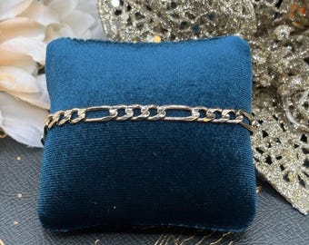 May include: A gold-colored Figaro chain bracelet displayed on a dark teal velvet cushion. The bracelet features a pattern of three small circular links followed by one elongated oval link, repeating along its length. The background includes decorative elements.