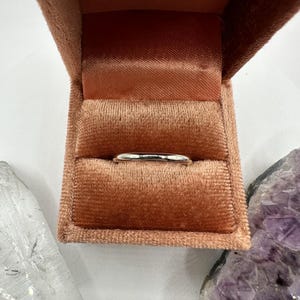 May include: A simple silver band ring in a velvet ring box. The box is a light peach color.