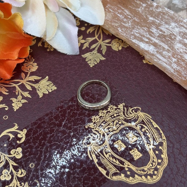 May include: A silver ring with small, clear stones set around the band. The ring is on a dark burgundy surface with gold floral and crest designs, and a few flowers in the background.