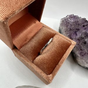 May include: A silver ring in a peach-colored velvet ring box. The box is open and the ring is sitting inside. The box is next to a large amethyst crystal.