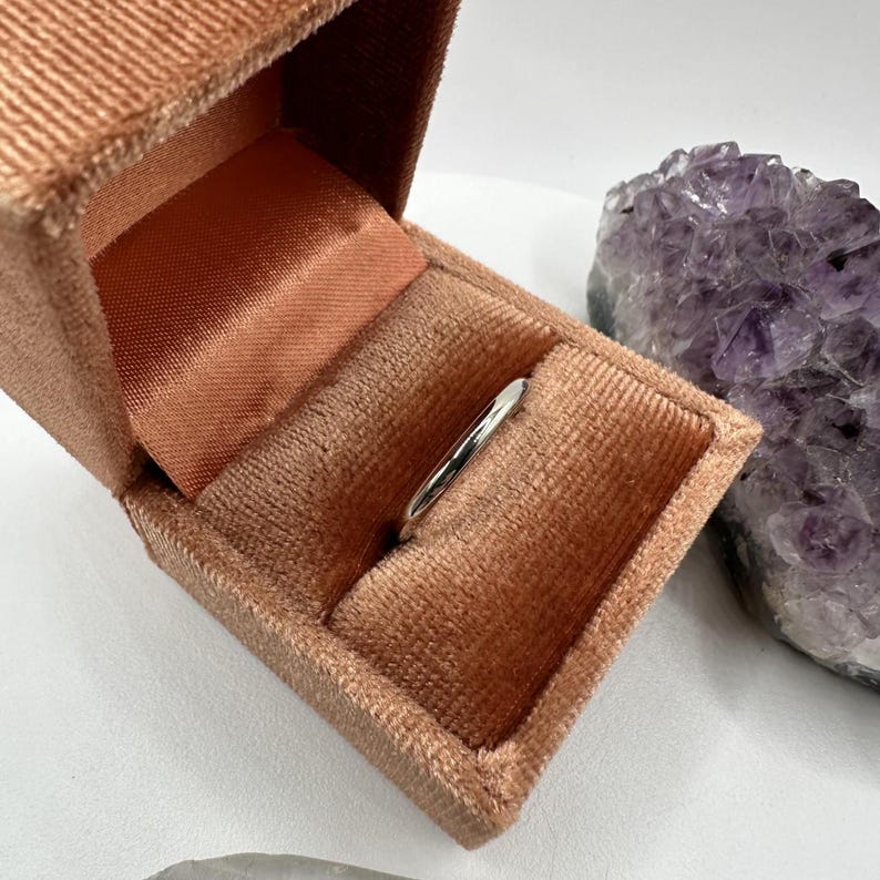 May include: A silver ring in a peach-colored velvet ring box. The box is open and the ring is sitting inside. The box is next to a large amethyst crystal.