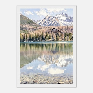 May include: A landscape print depicting a tranquil lake reflecting snow-covered mountains and a tree-lined shore. A large building is situated amongst the trees. The water is clear, showing the lakebed. The sky is a pale blue with fluffy clouds.