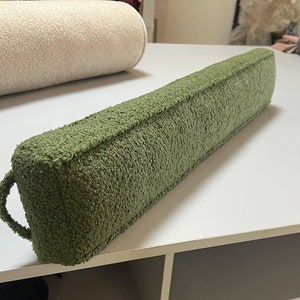 May include: A long, green, fuzzy doorstop with a loop handle. The doorstop is made of a soft, plush material and is designed to prevent doors from slamming.