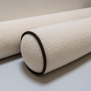 Peut inclure: Deux coussins cylindriques crème avec une surface texturée et moelleuse. Chaque coussin a une bordure marron foncé autour des extrémités arrondies. Les coussins sont posés sur une surface gris clair.