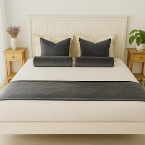 May include: A bed is made with a white mattress and headboard. It is decorated with gray velvet pillows, bolsters, and a bed runner. Two small wooden nightstands flank the bed, one with a vase of flowers and the other with a potted plant.