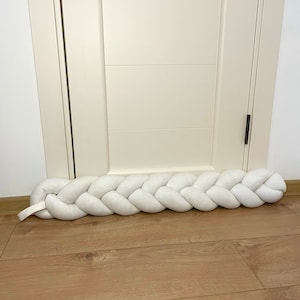 May include: A white braided doorstop made of fabric, placed at the bottom of a white door.