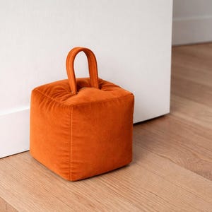 Puede incluir: Un tope de puerta cuadrado de terciopelo naranja, con un asa, colocado sobre un suelo de madera junto a un marco de puerta blanco. Este tope de puerta decorativo, un accesorio para el hogar, mide unos 15 cm de alto y ancho.