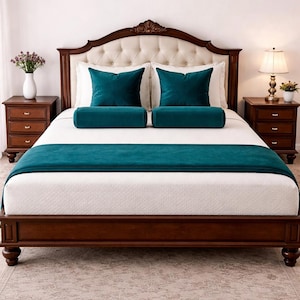 May include: A bed with a dark wood frame and headboard. The headboard is upholstered in a cream-colored fabric with button tufting. The bed is made with white bedding, teal pillows, and a teal bed runner. Two wooden nightstands flank the bed.