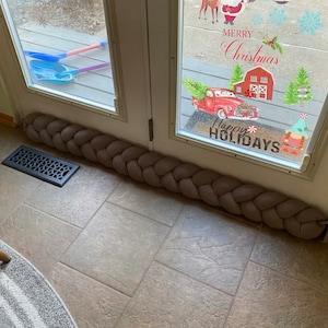 May include: A braided fabric door draft excluder in a neutral taupe colour. The long, cylindrical design is placed at the base of a glass door. The door has holiday-themed window clings.