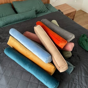 May include: A collection of cylindrical bolster pillows in various colors including teal, mustard yellow, light blue, dusty rose, beige, bright orange, grey, and olive green. Each pillow features a smooth velvet-like texture and a small fabric handle.