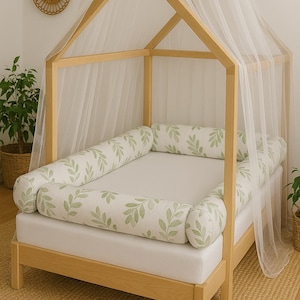 Puede incluir: Cama de bebé con estructura de madera y dosel. La cama tiene un parachoques con estampado de hojas y un dosel blanco. La cama está sobre una alfombra tejida. La estructura de la cama es de madera clara.