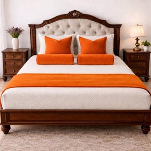 May include: A bed with a dark wood frame and headboard. The headboard is upholstered in a light beige fabric with a button-tufted design. The bed is made with white bedding, orange pillows, and a matching bed runner. Two wooden nightstands flank the bed.