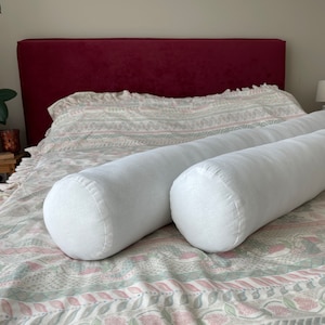 Könnte beinhalten: Zwei weiße zylindrische Kissen auf einem gemusterten Bettbezug. Der Bettbezug hat ein geometrisches Muster in Rosa, Grün und Weiß. Das Kopfteil ist dunkelrot.