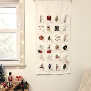 May include: A white fabric advent calendar with 24 pockets, each embroidered with a different Christmas-themed image and number. The calendar hangs from a wooden rod and is decorated with a garland.
