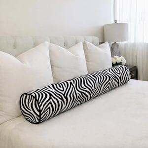May include: A bolster cushion with a black and white zebra print. The cushion is on a white bed, with white pillows and a buttoned headboard. A lamp and flowers are in the background. The cushion is approximately 75cm long.