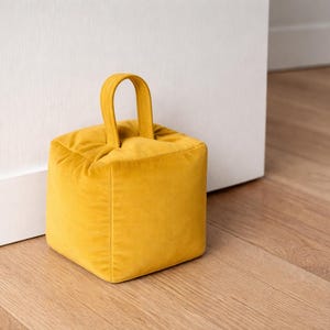 May include: A square, mustard-yellow doorstop made of soft velvet fabric. It features a small, arched handle on top for easy carrying. The doorstop is placed on a wooden floor next to a white door, providing a pop of color.