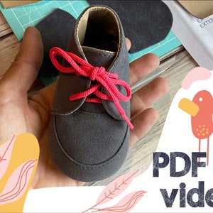 May include: A close-up of a gray baby shoe with pink laces. The shoe is being held in a hand. The background includes a piece of fabric with the words "Externo Gamuza" and a pattern of two birds.