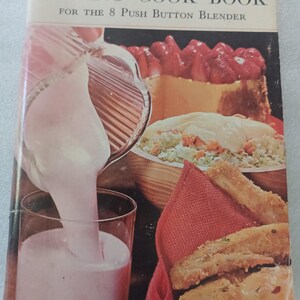 May include: Vintage cookbook titled "Waring Cook Book for the 8 Push Button Blender." The cover features a pink milkshake being poured into a glass, with images of food including a cake with strawberries, and a bowl of salad.